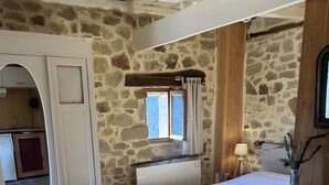Un dormitorio con pared de piedra, una cama, un espejo, una ventana y una pequeña mesa con un hervidor.