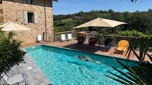 Una piscina con sillas de descanso y sombrillas, rodeada de edificios de piedra y vegetación.