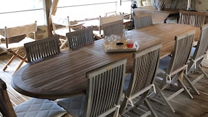 An outdoor wooden dining area with a high ceiling and exposed beams, surrounded by white cushioned chairs and a large table.