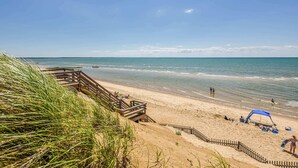 A beach with a wooden walkway leading to the shore, a sandy beach area with people and a blue canopy, and the ocean in the background. - Minutes from Beaches, Outdoor Shower, Fire Pit! (Eastham)