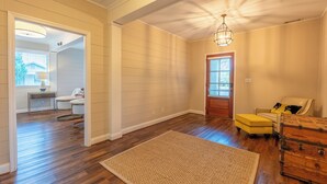 Una habitación amplia con pisos de madera, una alfombra grande, un reposapiés amarillo, un cofre de madera y una puerta que da a otra habitación.