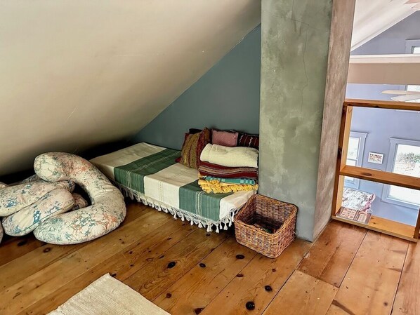 Une chambre mansardée chaleureuse avec un plafond en pente, un lit recouvert de plusieurs couvertures, un coussin décoratif, un panier en osier et une vue par la fenêtre.