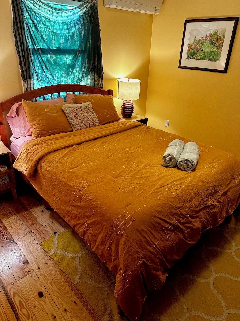 Una habitación con cama, lámpara de noche, un cuadro enmarcado en la pared y una ventana con cortina.