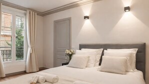 A neatly made bed with pillows and rolled towels, a window with curtains, and a wall-mounted light fixture. - 1525 Clef (Paris)