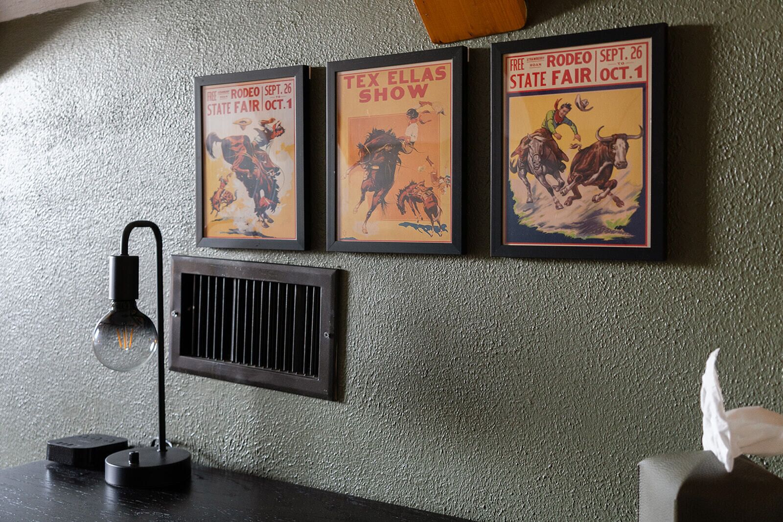 Tre poster incorniciati su una parete strutturata, una bocchetta di ventilazione nera e una lampada da scrivania.
