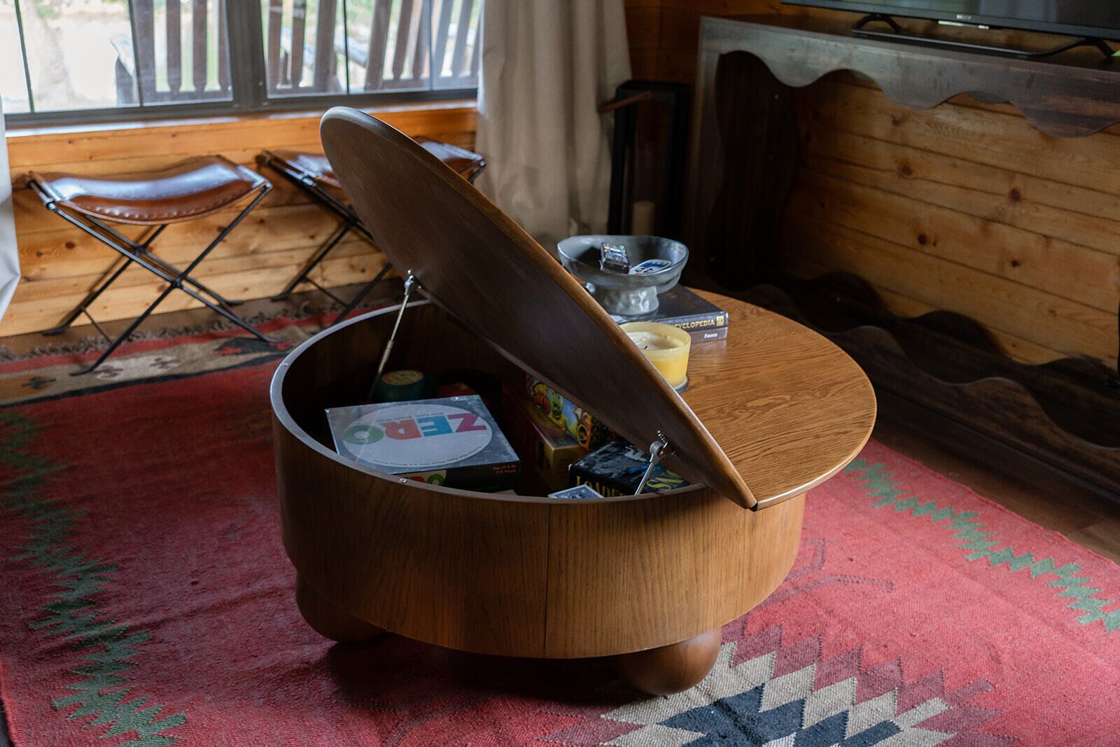 Une table basse en bois avec un couvercle, un tapis rouge à motifs et une fenêtre avec des rideaux en arrière-plan.