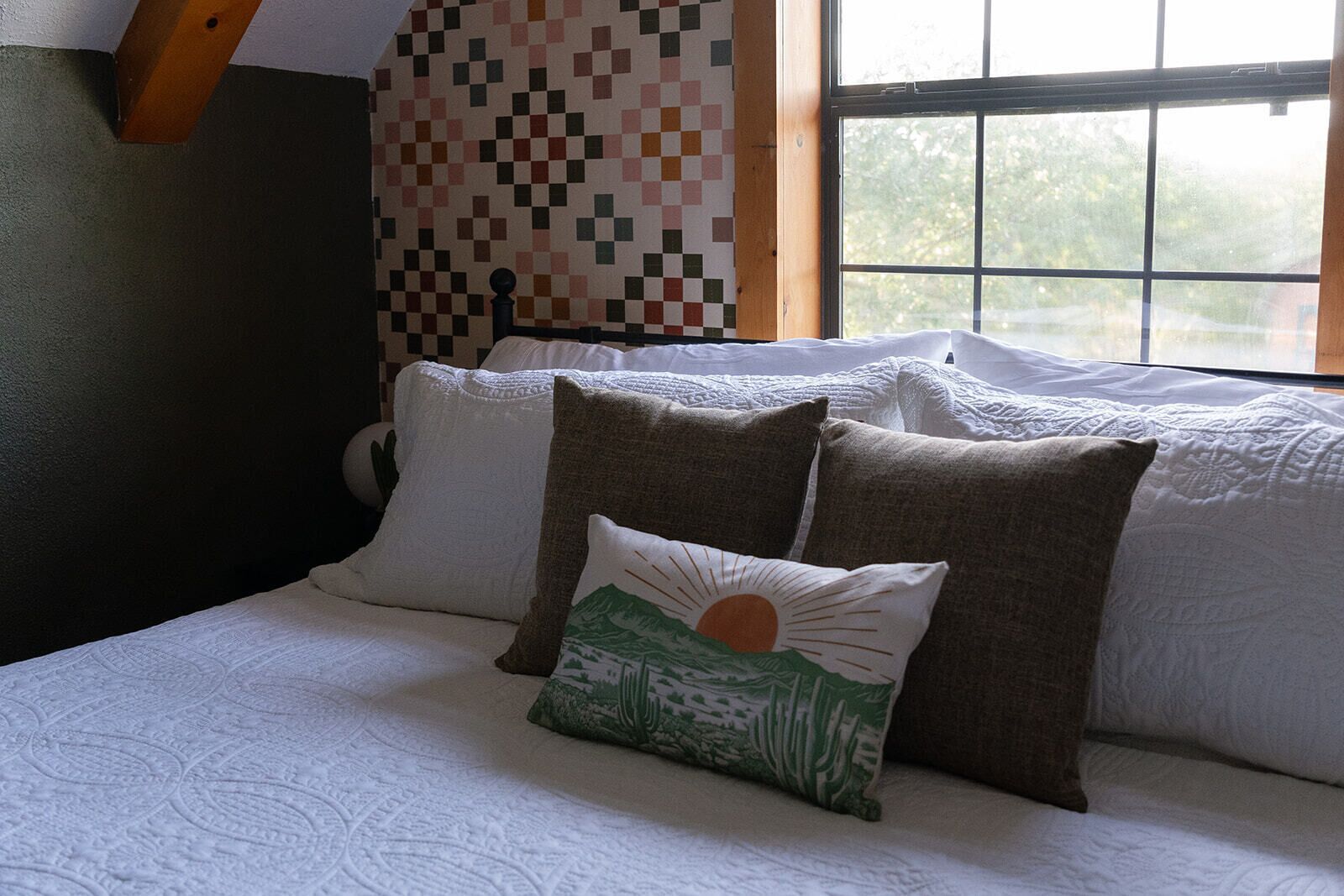 Un lit avec une couette blanche et plusieurs oreillers, dont un vert orné d’un motif représentant un paysage désertique.