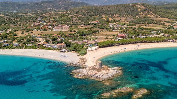 Vista aérea de un pueblo costero con playa, afloramientos rocosos y colinas circundantes.