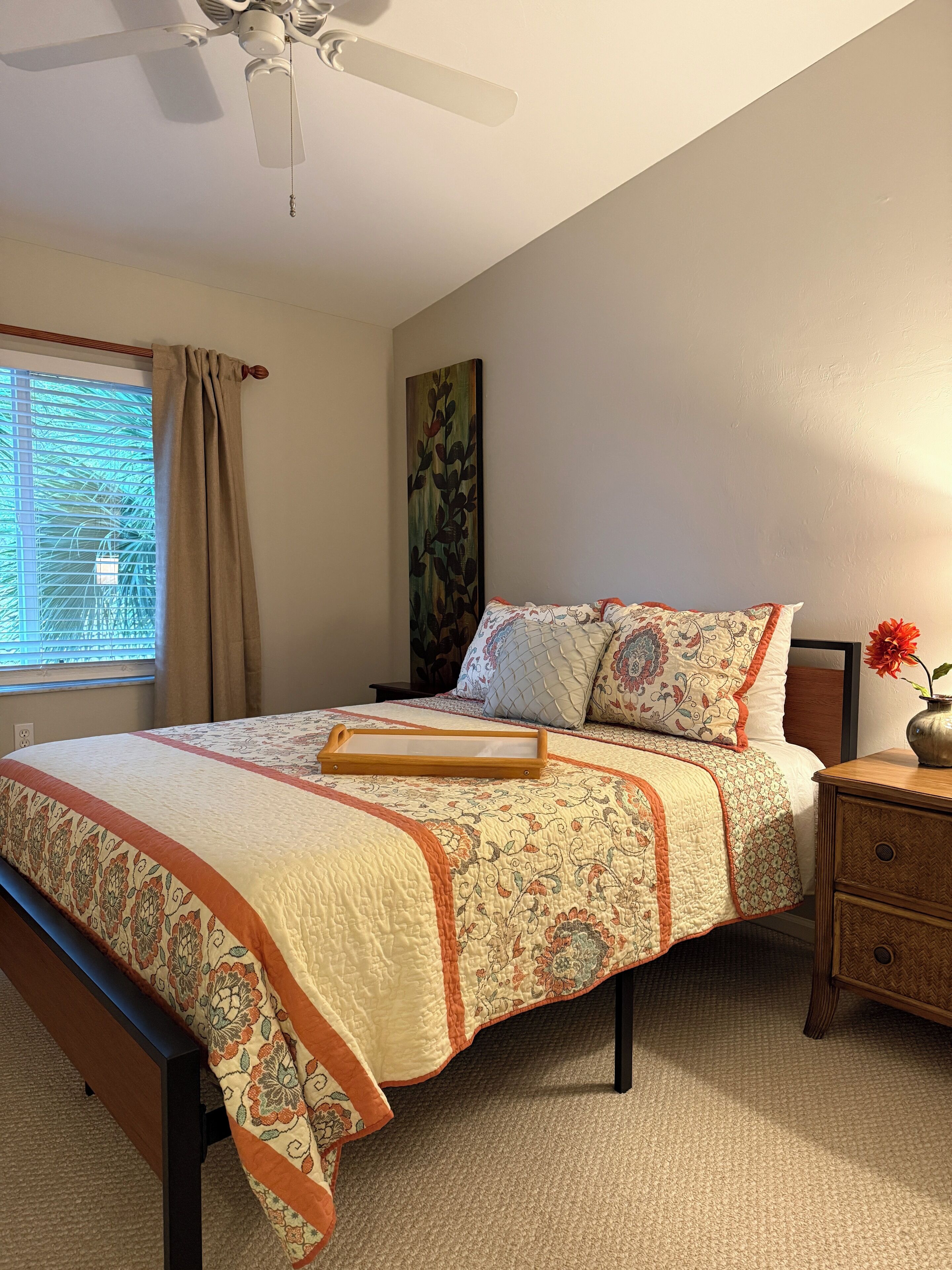 A bedroom with a bed, a nightstand, a vase with flowers, a ceiling fan, and a window with curtains.