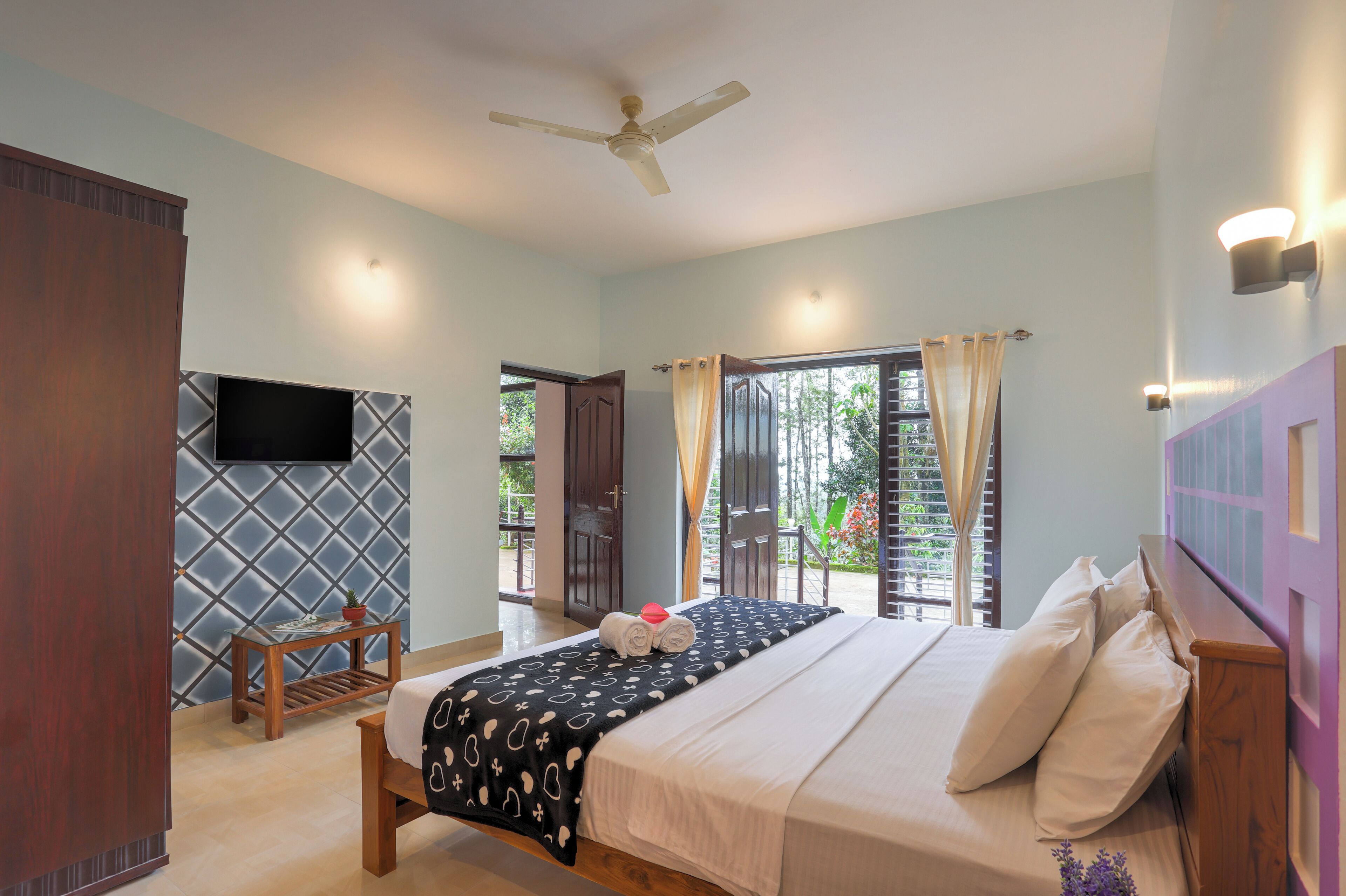 A bedroom with a bed, a ceiling fan, a TV mounted on the wall, and a window with curtains.