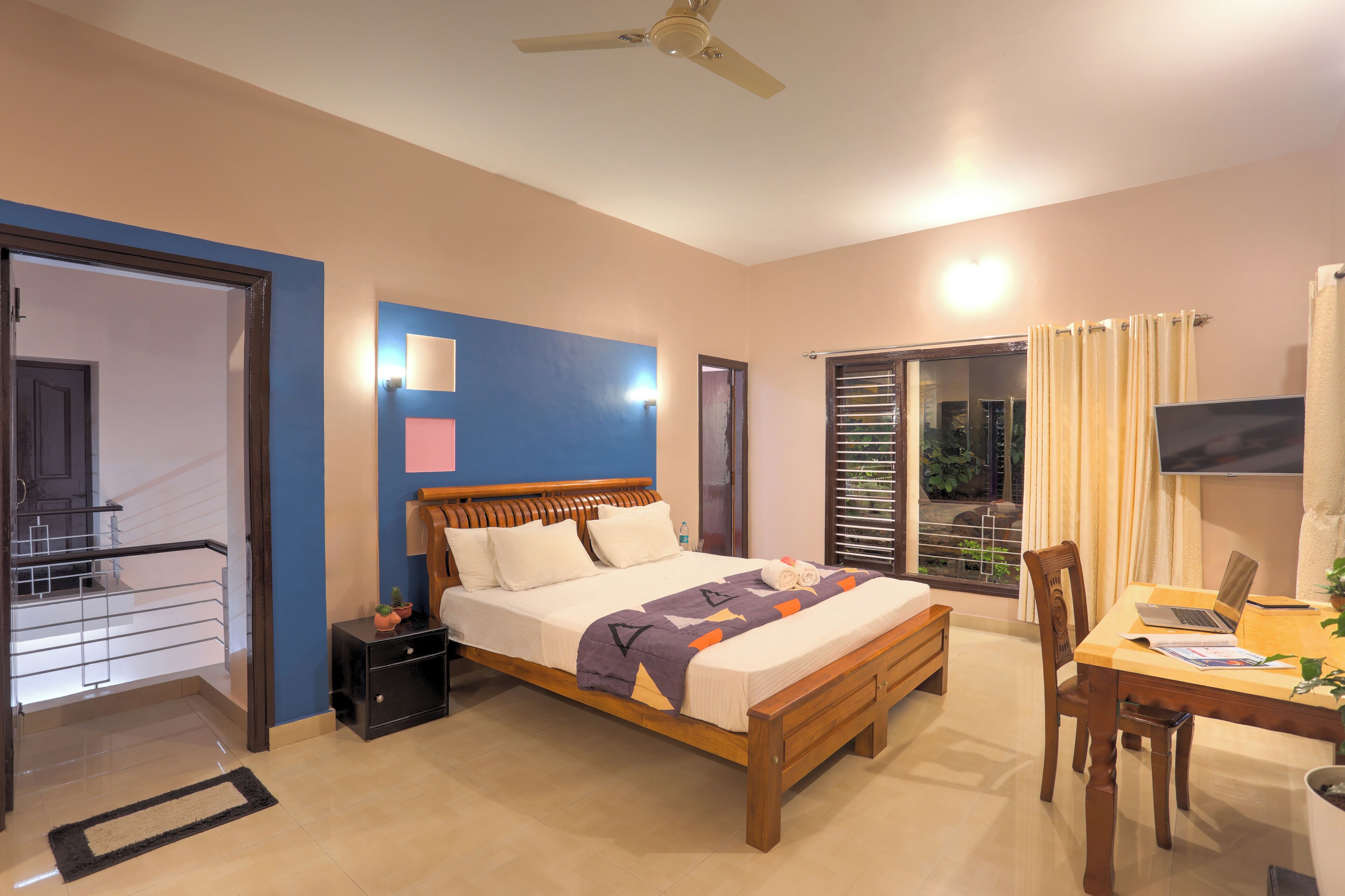 A hotel room with a bed, a desk with a laptop, a TV, and a balcony with plants.