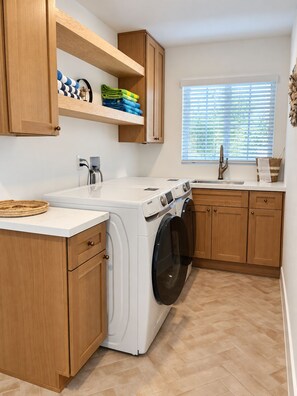 A laundry room with a front-loading washing machine, wooden cabinets, a window with blinds, and a sink. - Stunning 4BR/3BA Home in Sunset Cliffs – Walk to Ocean (San Diego)