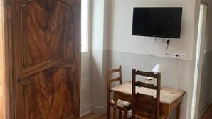 A room with a wooden cabinet, a dining table with chairs, a flat-screen TV mounted on the wall, and a window with curtains. - Au Sans Souci (Saint-Paul-lès-Monestier)