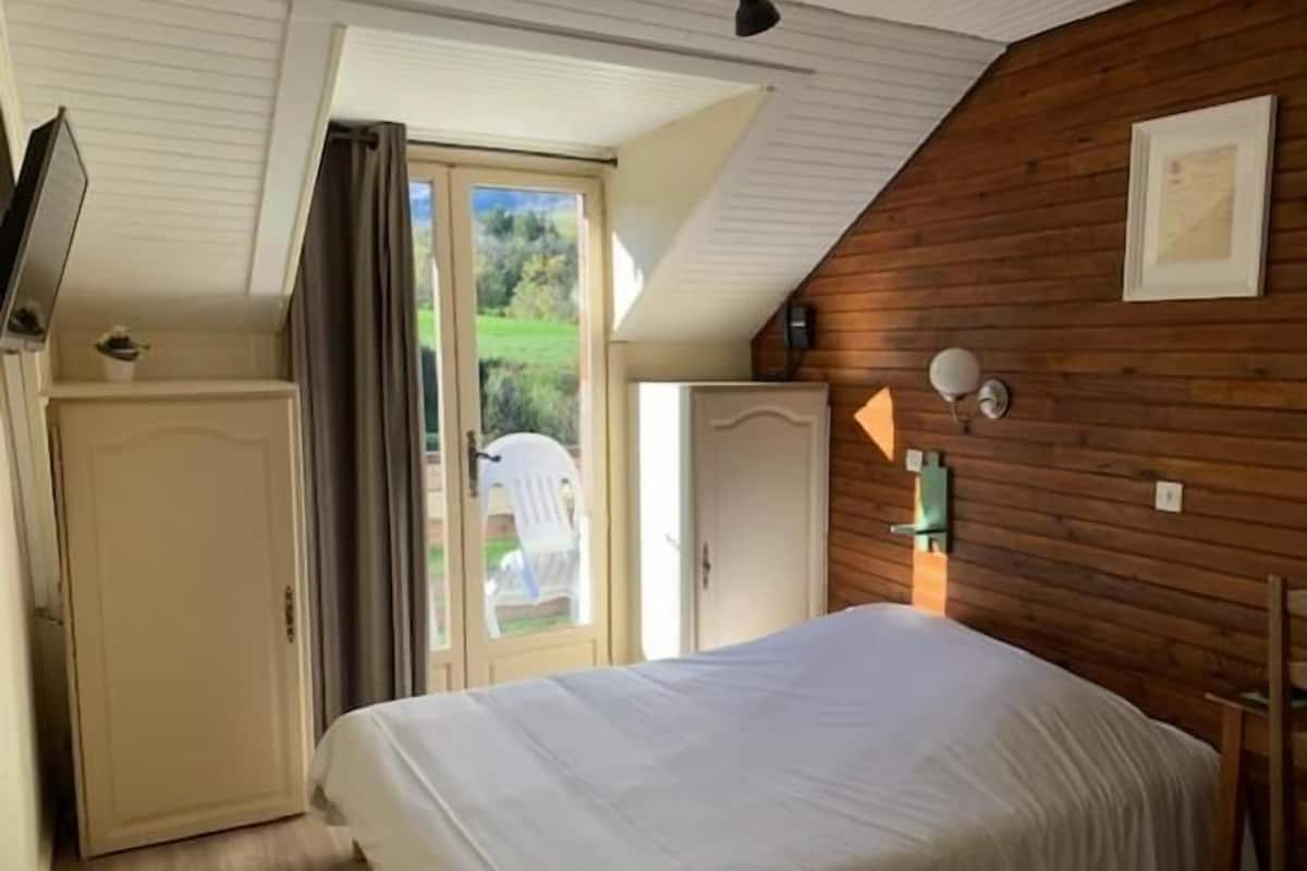 Een slaapkamer met een bed, een kledingkast, een deur en een raam met uitzicht op groen.
