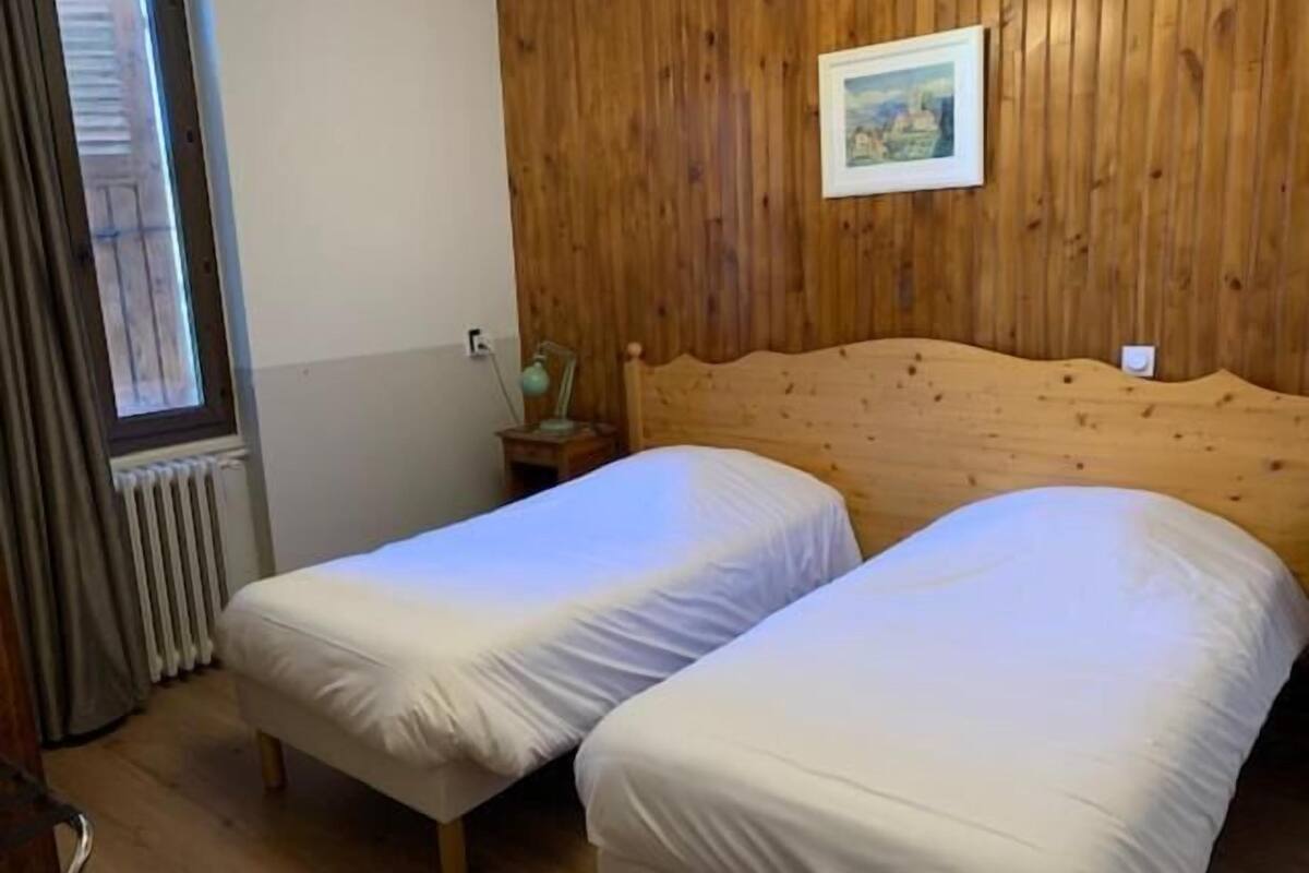 Een kamer met twee eenpersoonsbedden, houten wanden, een klein nachtkastje met een lamp, een raam met gordijnen en een radiator.