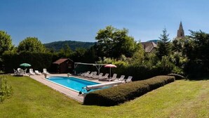 A swimming pool with lounge chairs and umbrellas, surrounded by well-maintained grass and trees, with a building and a church in the background. - Au Sans Souci (Saint-Paul-lès-Monestier)