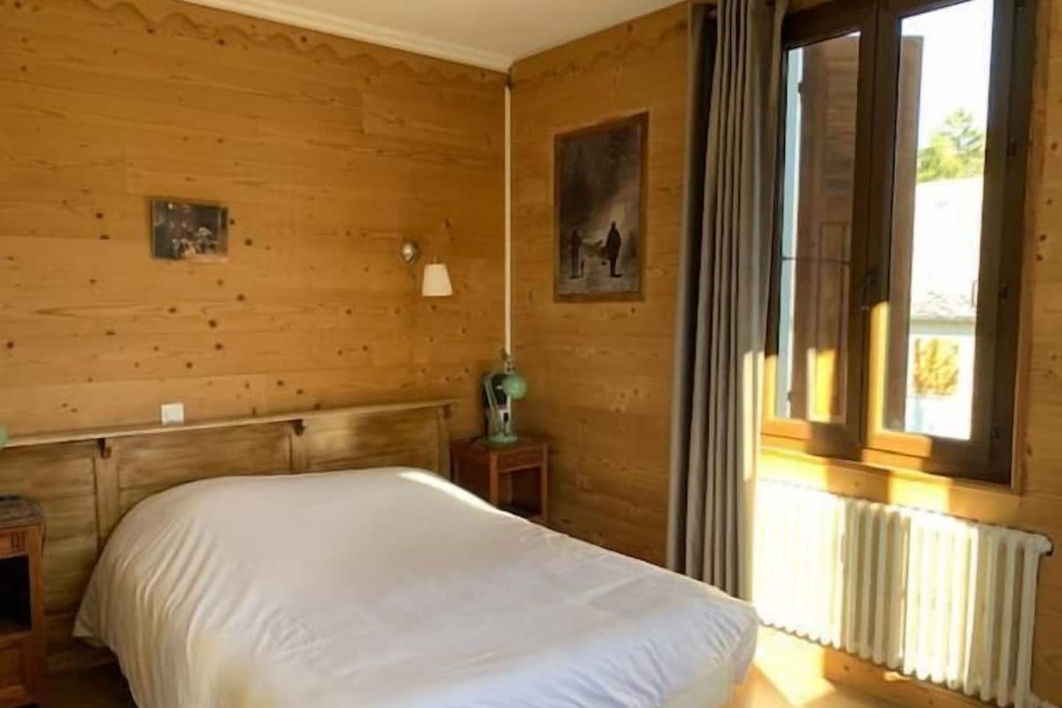 Een kamer met houten lambrisering, een bed, een klein tafeltje met een lamp en een raam met gordijnen.