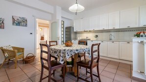 Comedor con mesa redonda, cuatro sillas, lámpara colgante y cocina con armarios blancos y salpicadero de azulejos.