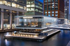 A modern building with a glass facade, named "Hawksmoor," featuring a rooftop bar and a waterfront patio. - Modern 2-Bedroom Riverside Apartment Near Canary Wharf (London)