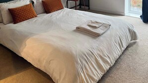 Een netjes opgemaakt bed met witte lakens en bruine kussens, een klein bureau met een stoel en een raam met uitzicht op het omliggende landschap.