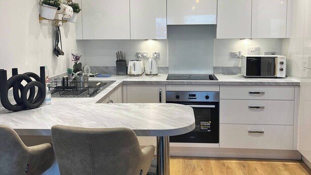 Een moderne keuken met witte kasten, een ingebouwde oven en een magnetron. Er is een eethoek met een marmeren tafel en twee stoelen.