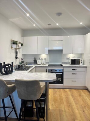 A modern kitchen with white cabinets, a built-in oven, and a microwave. There is a dining area with a marble table and two chairs. - Luxury Apartment - Lakeside (Grays)