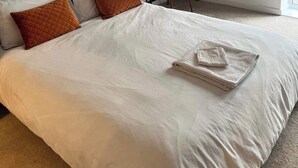 Een net opgemaakt bed met witte lakens en twee bruine kussens, een klein bureau met stoel, en een schilderij aan de muur.
