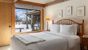 Ein Schlafzimmer mit großem Fenster, Holzbalken, einem Bett mit weißer Bettwäsche, einem Nachttisch mit Lampe und gerahmten floralen Bildern an der Wand.