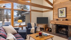 Ein gemütliches Wohnzimmer mit Kamin, Fernseher und einem großen Fenster mit Blick auf eine verschneite Landschaft.