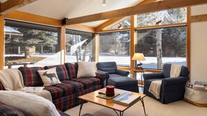 Ein gemütliches Wohnzimmer mit einem karierten Sofa, Sesseln und einem Couchtisch. Große Fenster bieten Blick ins Freie.