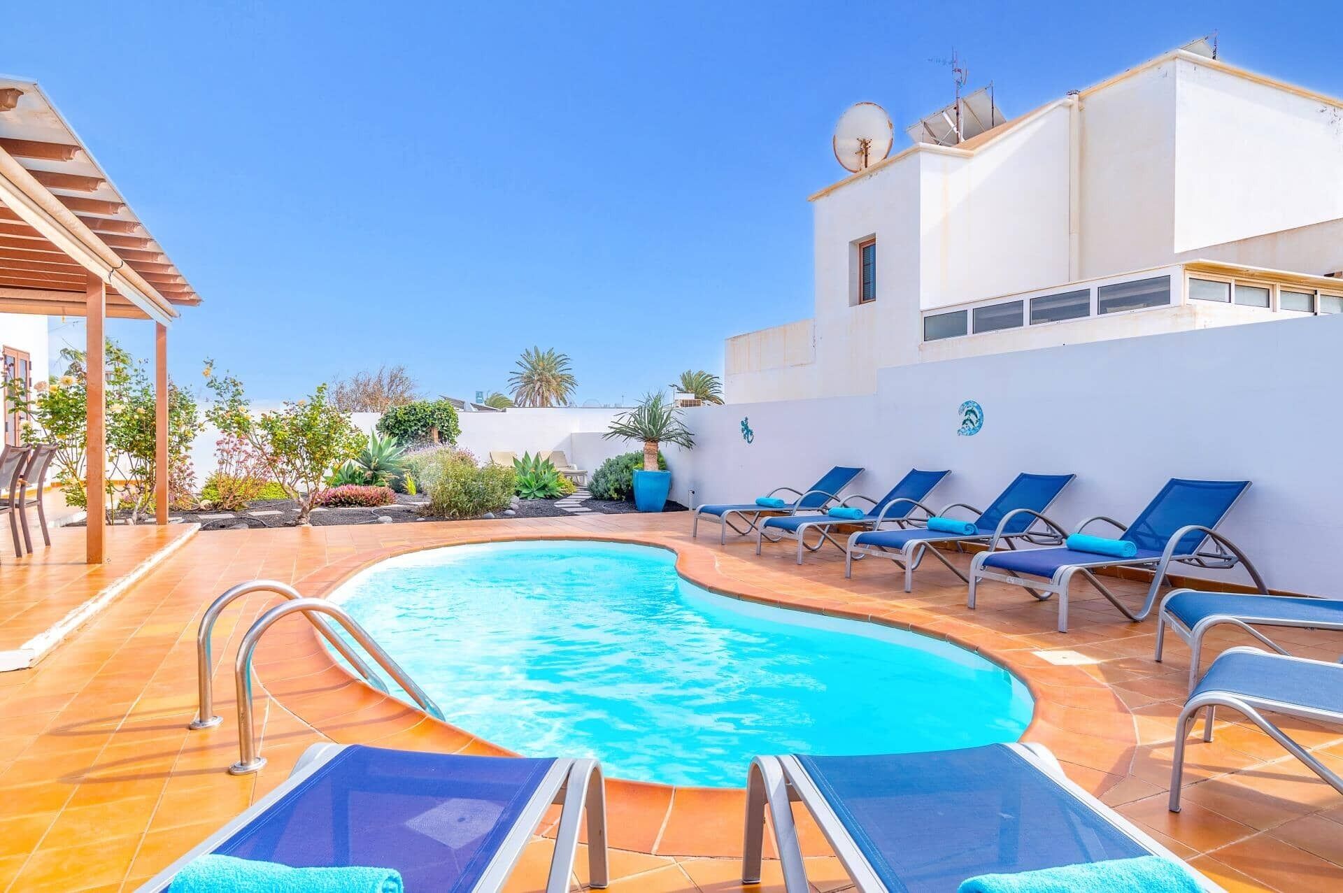 Une piscine dotée de chaises longues et d’une pergola, entourée d’un bâtiment blanc sur lequel est installée une antenne parabolique.