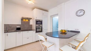 Eine moderne Küche mit weißen Schränken, einem Backofen, einer Mikrowelle und einem Spülbecken. Zwei weiße Stühle mit Holzbeinen stehen an einer gewölbten Kochinsel. Eine Uhr ist an der Wand befestigt.