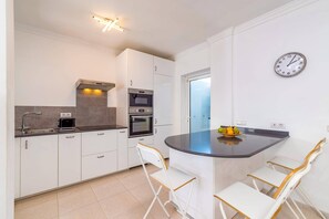Eine moderne Küche mit weißen Schränken, einem Backofen, einer Mikrowelle und einem Spülbecken. Zwei weiße Stühle mit Holzbeinen stehen an einer gewölbten Kochinsel. Eine Uhr ist an der Wand befestigt.