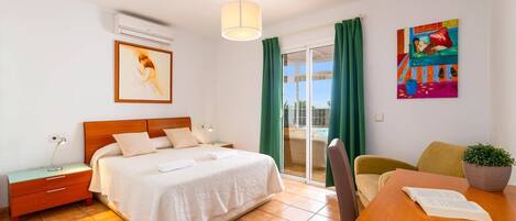 Een hotelkamer met een bed, een bureau, een stoel, een bijzettafeltje, een nachtkastje, een airconditioner, een raam met gordijnen en een schilderij aan de muur.
