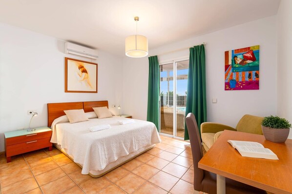A hotel room with a bed, a desk, a chair, a small table, a nightstand, an air conditioning unit, a window with curtains, and a painting on the wall. - Villa Papagayo Faro by Villa Plus (Playa Blanca)