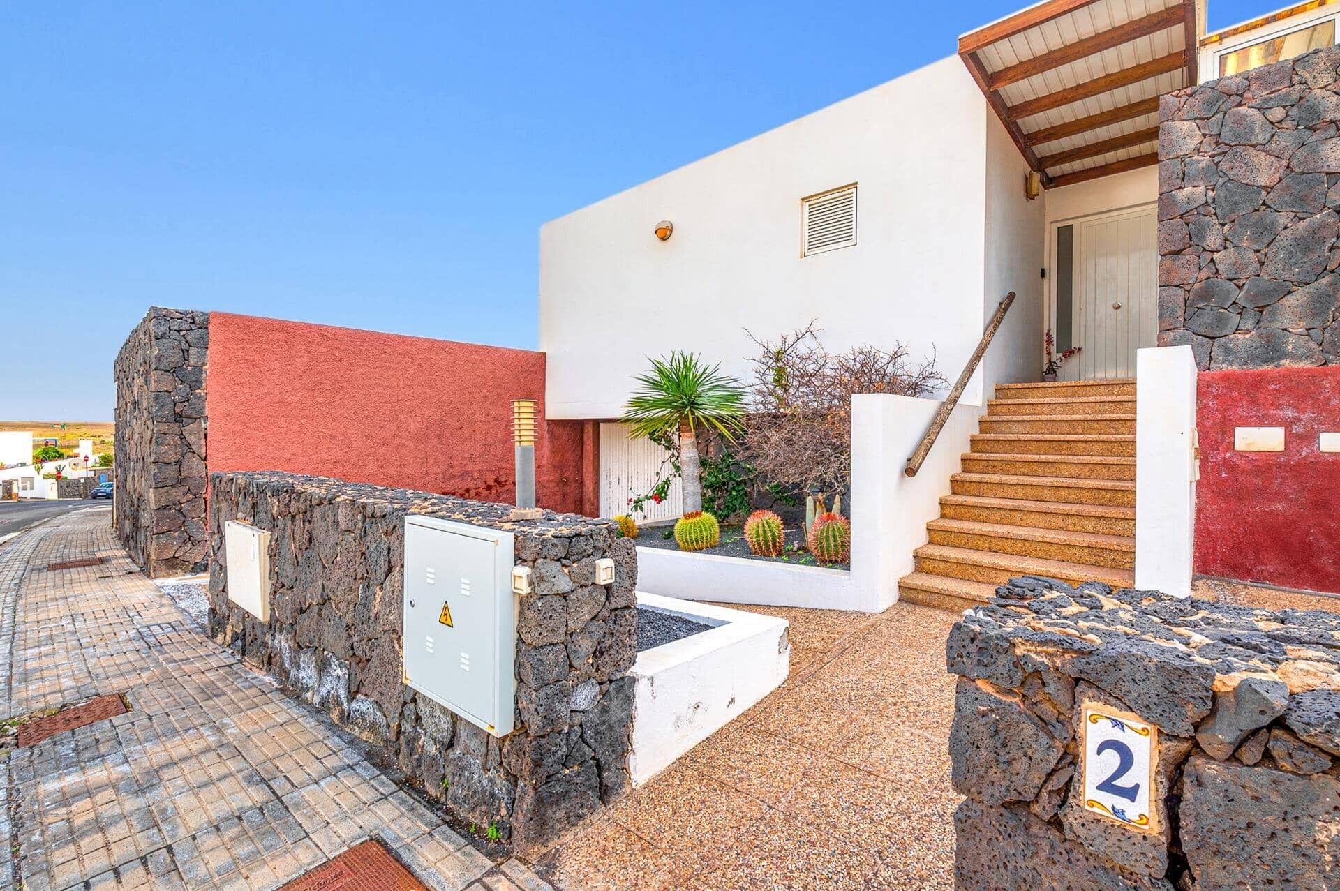 A building with a white facade, stone walls, and a red door. There is a staircase leading to a covered entrance, and a small garden with cacti and a palm tree.