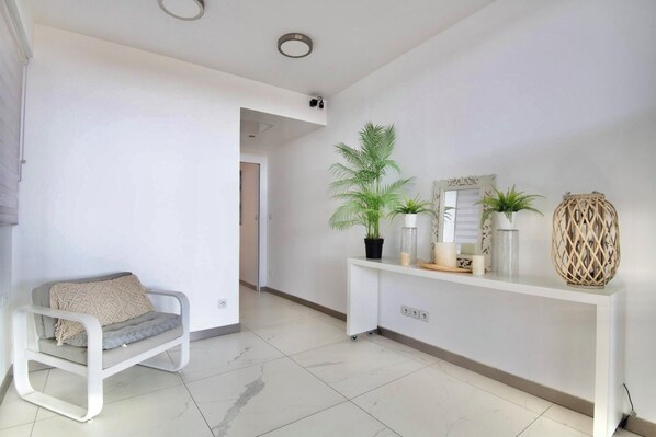 Un salon moderne avec un canapé blanc, une console ornée d’objets décoratifs et un couloir menant à une autre pièce.