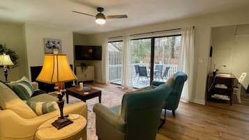 Un salon comprenant un canapé, des fauteuils, une table basse, une lampe, un téléviseur et une porte-fenêtre coulissante donnant sur un balcon.