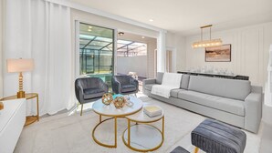 A modern living room with a grey sofa, two armchairs, a coffee table, and a dining area with a chandelier. - Modern 5BR Home w/ Pool at Windsor Island (Davenport)
