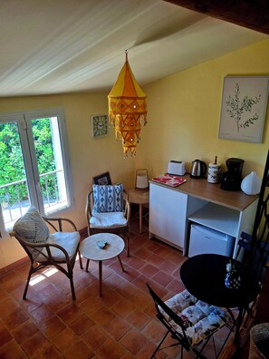 A cozy room with a wicker chair, a small round table, a hanging light fixture, a white refrigerator, and a window with a view of greenery. - Guest rooms adjoining the main house with a separate entrance (Bédoin)