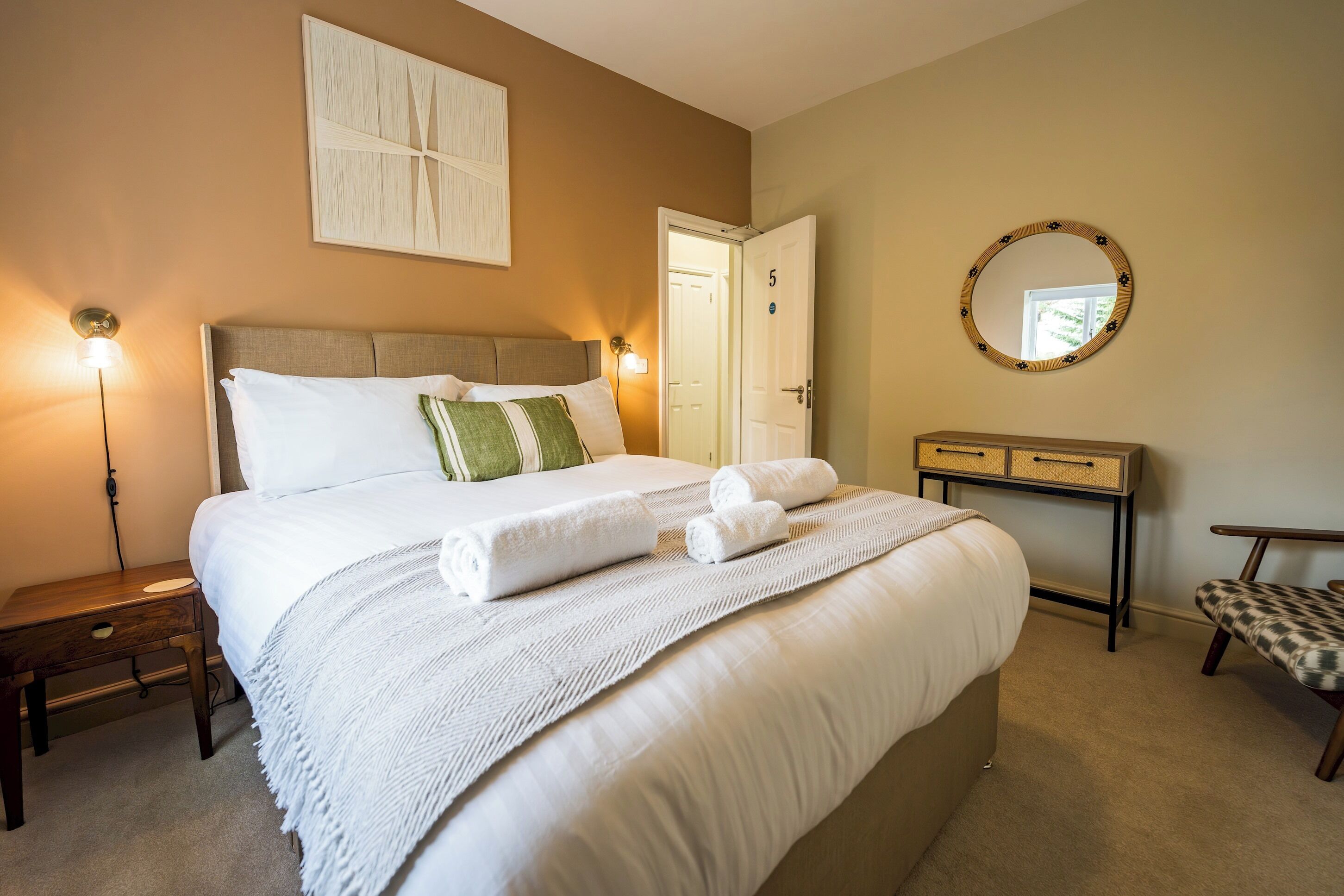 A neatly made bed with white linens and a green pillow, a bedside table with a lamp, a round mirror on the wall, and a door with the number 5.