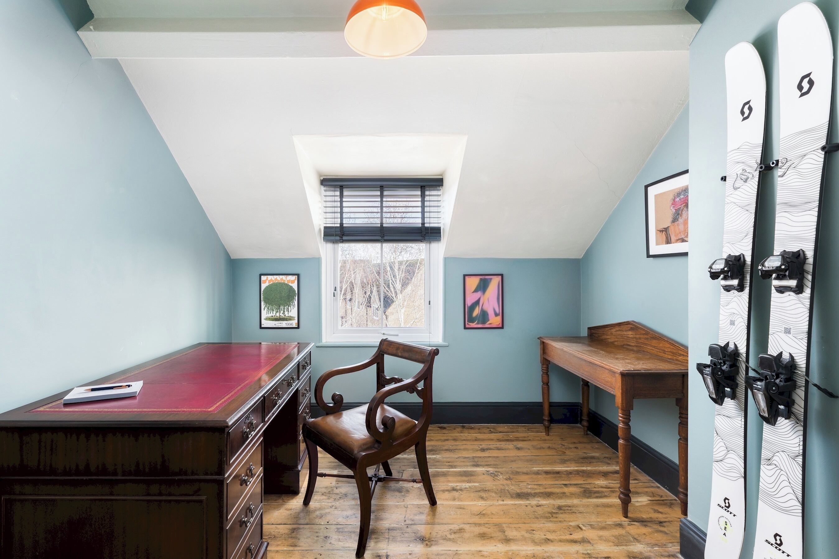 A room with a sloped ceiling, a wooden desk, a chair, a window with blinds, and skis mounted on the wall.
