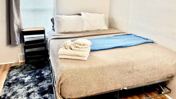 A small, neatly arranged bedroom with two beds, a window with blinds, a small table, and a rug. - 3-bedroom house ~ Pro Cleaned - Work & Family Friendly (Racine)