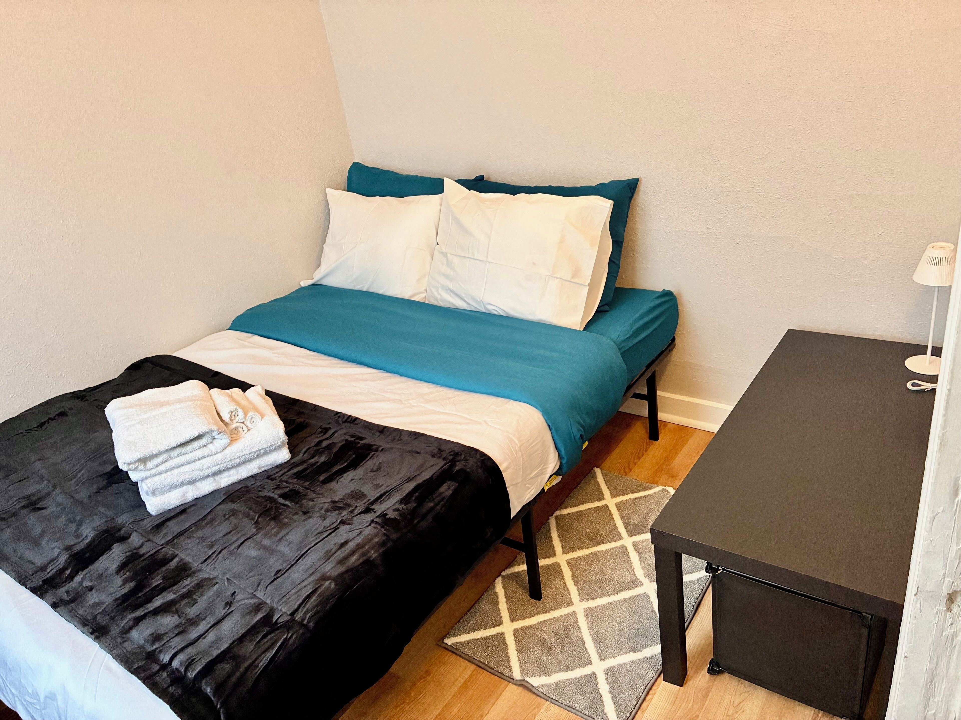 A neatly made bed with a black and teal bedspread, white pillows, and a folded towel on the left side. There is a small table with a lamp beside the bed, and a patterned rug on the floor.