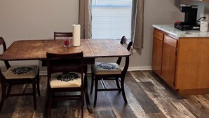 A dining area with a wooden table and chairs, a window with blinds, and a kitchen area with wooden cabinets and a coffee maker. - 3-bedroom house ~ Pro Cleaned - Work & Family Friendly (Racine)