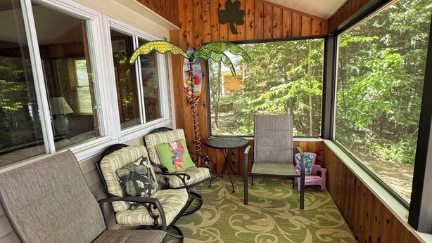 A covered porch with wooden walls, a green patterned carpet, and a view of the forest. - Lakefront Family Home with AC, Dock, Beach & Screened Deck (Harrison)