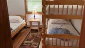 A room with two bunk beds, a small bedside table with a lamp, a window with a view, and a patterned rug on the floor. - Lakefront Family Home with AC, Dock, Beach & Screened Deck (Harrison)