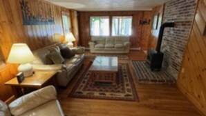 A cozy living room with wooden walls, a sofa, a coffee table, and a fireplace. - Lakefront Family Home with AC, Dock, Beach & Screened Deck (Harrison)