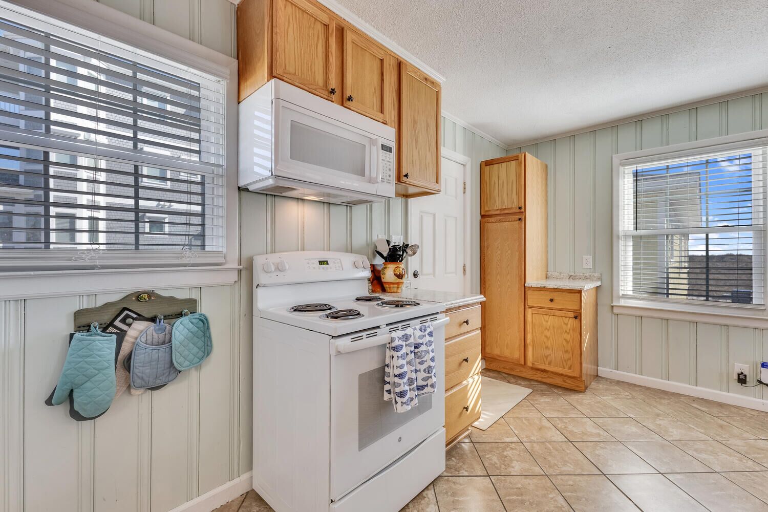 Una cocina con armarios de madera, una estufa blanca y un microondas. Hay dos ventanas con persianas, y una puerta que conduce a otra habitación.