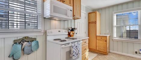 Una cocina con armarios de madera, una estufa blanca y un microondas. Hay dos ventanas con persianas, y una puerta que conduce a otra habitación.